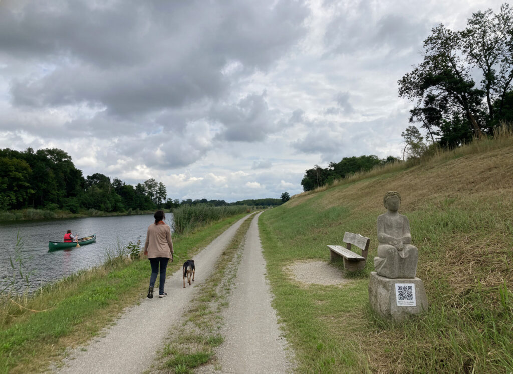 Beispielhafte Darstellung eines Kanufahrers und einer Spaziergängerin mit Hund am Elbe-Havel-Kanal, vorbei an einer Sitzbank und einer figürlichen Darstellung einer Person aus Stein.
